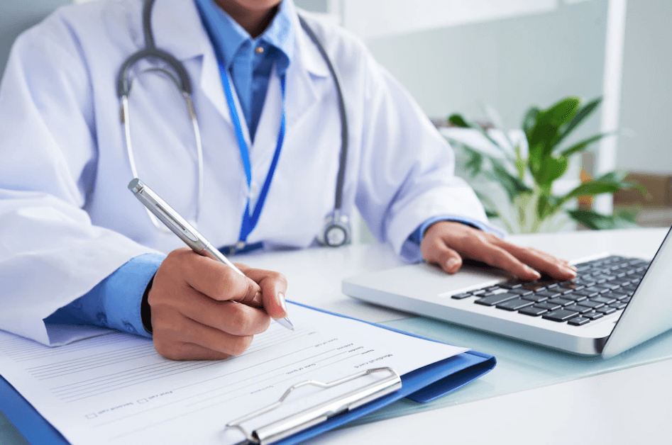 A female doctor with a stethoscope writing on a form while typing on a laptop keyboard, representing medical education for Pakistanis in the UK.